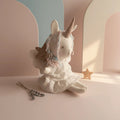 Muñeca unicornio de tela hecha a mano, con vestido blanco y crin en tonos pastel, sentada sobre fondo rosa pastel.