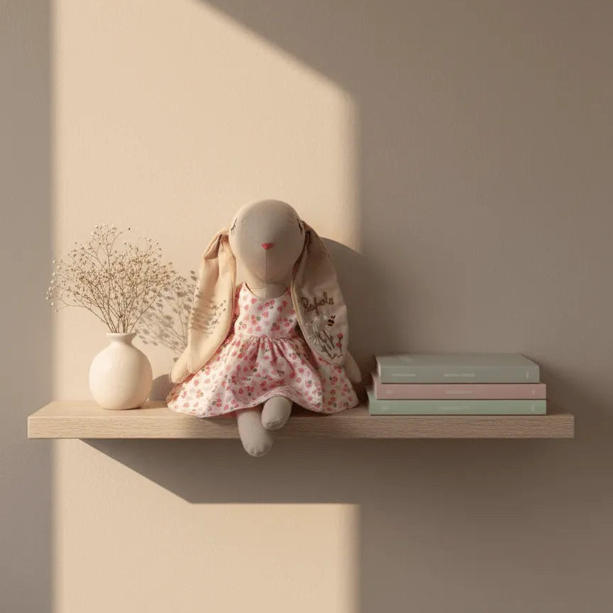 Conejita de tela hecha a mano con vestido floral rosa, sentada sobre una repisa de madera en habitación infantil de tonos neutros.