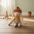 Conejito de tela artesanal con gorro terracota y cuello tejido mostaza, sentado en el suelo de una habitación infantil estilo nórdico.
