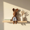 Conejito amigurumi café con peto celeste, tejido a mano, sentado sobre una repisa de madera en habitación infantil de tonos neutros.