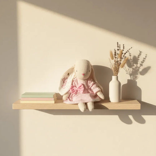 Conejita de peluche de tela con vestido rosa pastel y lazo grande, sentada sobre una repisa de madera en habitación infantil iluminada por el sol.