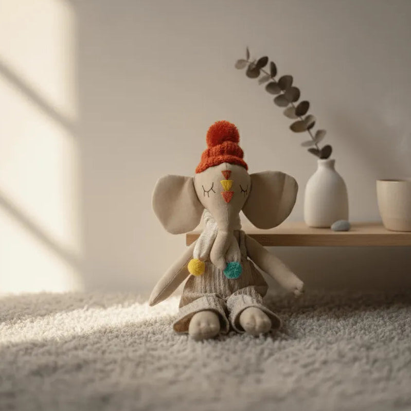 Muñeco de elefante de tela con gorro naranja y pompones, sentado sobre alfombra suave junto a una repisa con taza y jarrón decorativo.