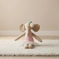 Muñeca de elefantita de tela con vestido rosa y falda de tul, sentada sobre una alfombra blanca en una habitación infantil de tonos neutros.