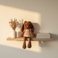 Conejita amigurumi café con vestido rosa pastel, sentada sobre una repisa de madera junto a libros y jarrón con flores secas.