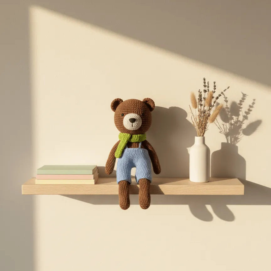 Osito amigurumi café con jardinera celeste y bufanda verde, sentado sobre una repisa de madera junto a libros y jarrón con flores secas.