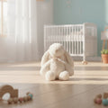 Conejito de peluche blanco extra suave con lazo de organza, sentado en el piso de una habitación de bebé con cuna blanca al fondo