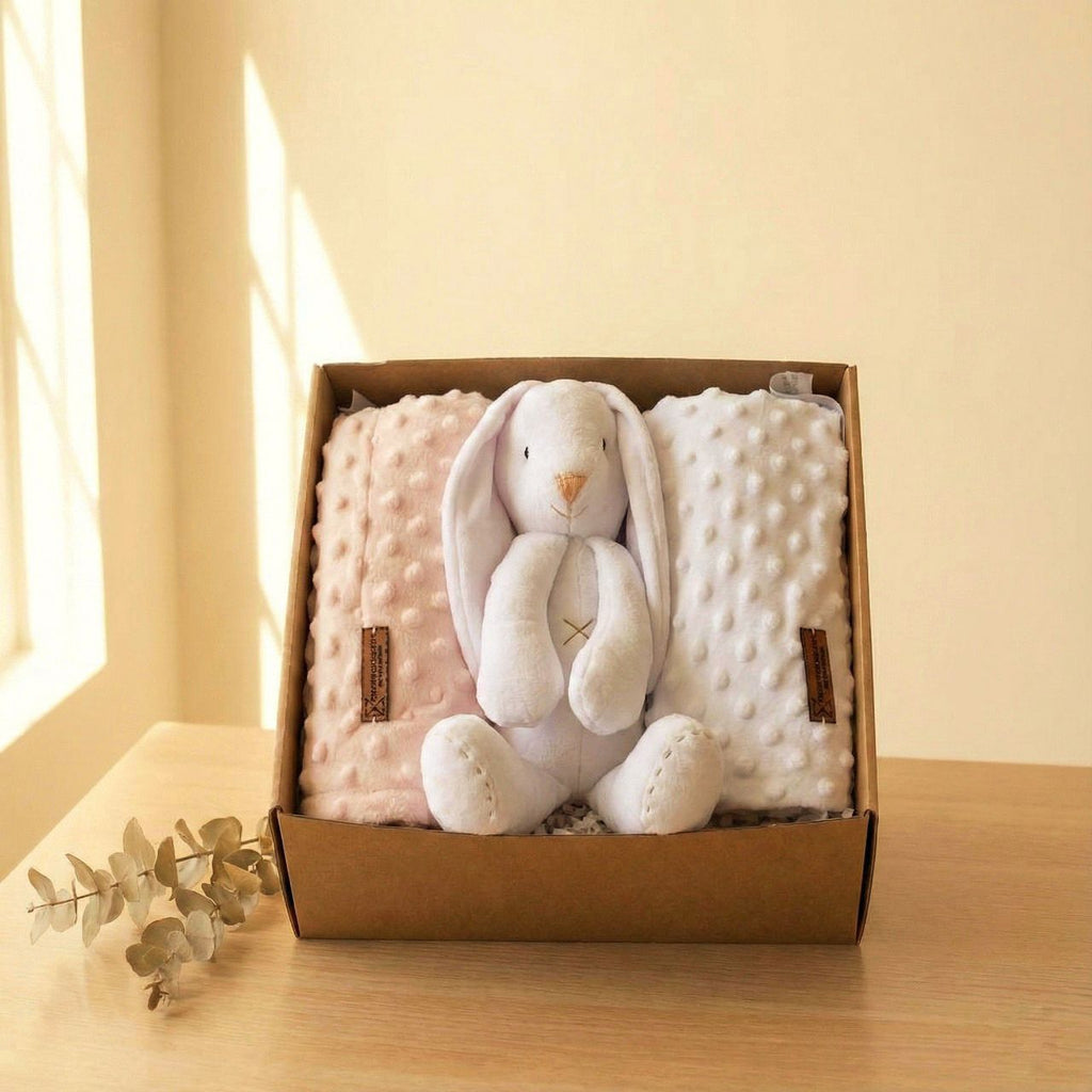 Set regalo para bebé con conejito de peluche blanco y dos mantas suaves rosa y blanca dentro de una caja kraft sobre mesa de madera.