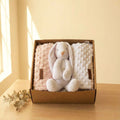 Set regalo para bebé con conejito de peluche blanco y dos mantas suaves rosa y blanca dentro de una caja kraft sobre mesa de madera.