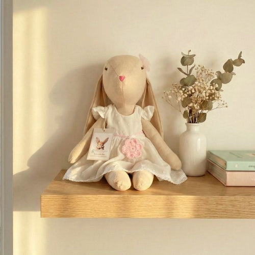 Conejita de tela color miel con vestido blanco y flor rosa, sentada sobre una repisa de madera junto a jarrón con flores secas y libros pastel.”