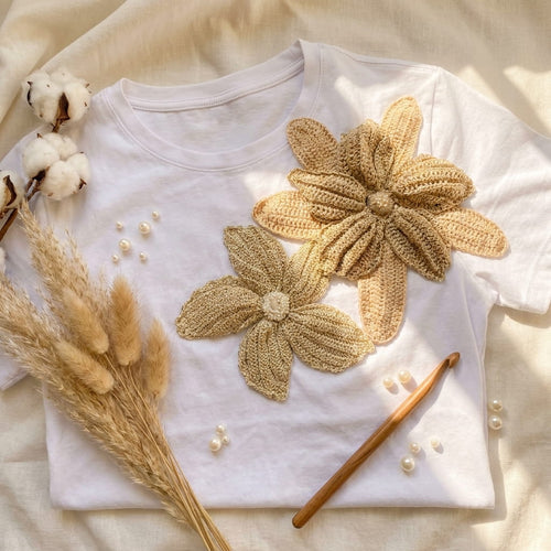 Camiseta artesanal colombiana con flores crochet 3D en tonos dorados, flat lay con pampa grass y perlas